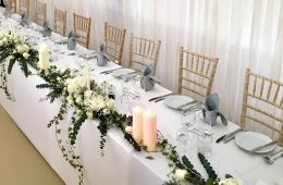 Bright white wedding top tables with grey napkins and green foliage runners with white roses and candles